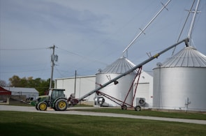 A green tractor is parked on a gravel path near a farm building. Adjacent to the tractor, there is an agricultural auger conveyor connected to large silver grain silos. Several power lines and a utility pole are visible, with grassy areas surrounding the scene.