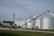 A green tractor is parked on a gravel path near a farm building. Adjacent to the tractor, there is an agricultural auger conveyor connected to large silver grain silos. Several power lines and a utility pole are visible, with grassy areas surrounding the scene.