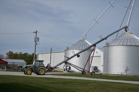 A green tractor is parked on a gravel path near a farm building. Adjacent to the tractor, there is an agricultural auger conveyor connected to large silver grain silos. Several power lines and a utility pole are visible, with grassy areas surrounding the scene.
