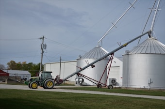 A green tractor is parked on a gravel path near a farm building. Adjacent to the tractor, there is an agricultural auger conveyor connected to large silver grain silos. Several power lines and a utility pole are visible, with grassy areas surrounding the scene.