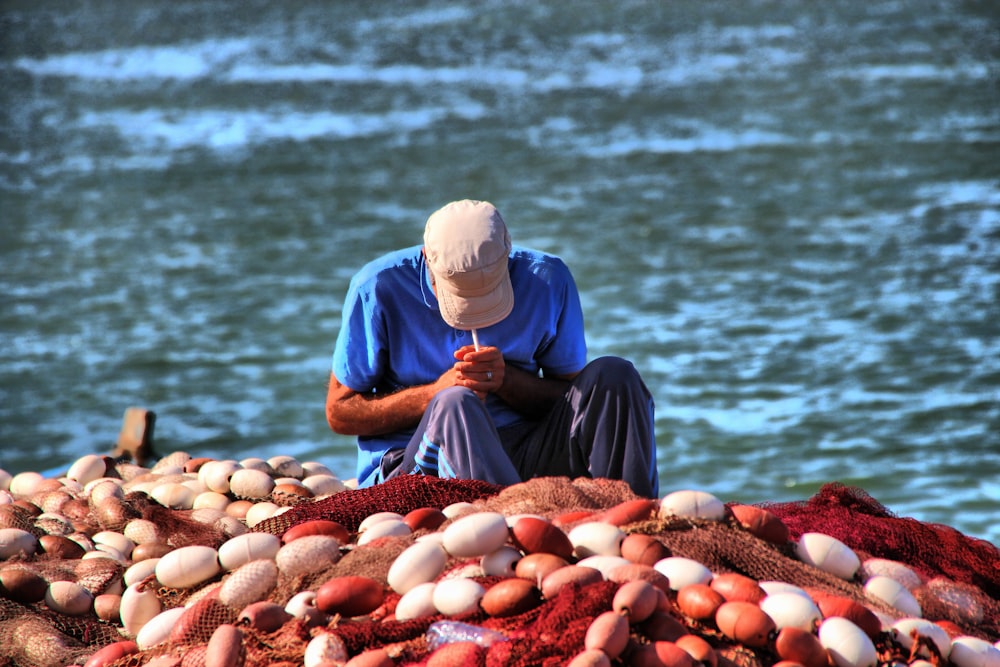 man in blue shirt and white cap sitting on brown rock near body of water during