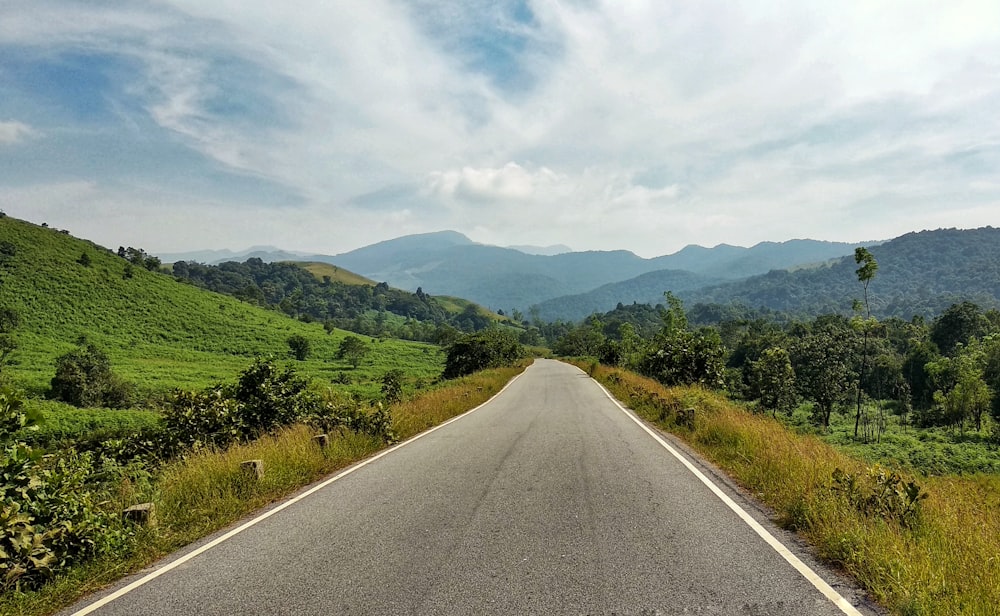 gray asphalt road between green grass field under white cloudy sky during daytime