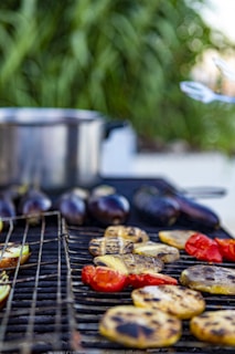 A close-up of a barbecue grill with various vegetables, including red bell peppers, eggplants, and sliced potatoes, being roasted. A silver pot is visible in the background, with a blurred backdrop of greenery.