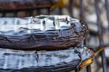 Charred and glossy vegetables, possibly eggplants, rest on a grill, their skin darkened and slightly wrinkled from grilling. The slick, shiny surface reflects light, highlighting the texture.