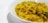 A close-up view of a bowl filled with yellow, curly noodles garnished with small pieces of green herbs and possibly some vegetables. The noodles have a shiny, cooked appearance, suggesting they are seasoned with spices.