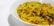 A close-up view of a bowl filled with yellow, curly noodles garnished with small pieces of green herbs and possibly some vegetables. The noodles have a shiny, cooked appearance, suggesting they are seasoned with spices.