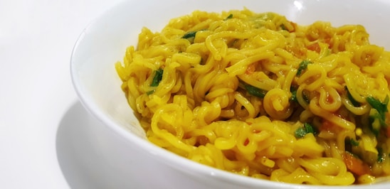 A close-up view of a bowl filled with yellow, curly noodles garnished with small pieces of green herbs and possibly some vegetables. The noodles have a shiny, cooked appearance, suggesting they are seasoned with spices.