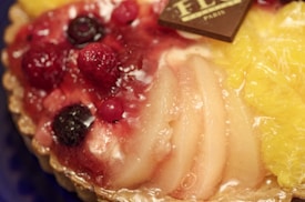 A close-up view of a fruit tart adorned with various fruits such as raspberries, blackberries, and orange slices. The surface is glossy, reflecting the syrupy glaze enhancing the vibrant colors. A small piece of chocolate with text engraved on it is placed on top, partly obscured by the fruits.