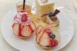 A variety of elegant desserts is arranged on a white plate. The selection includes pastries topped with strawberries, raspberries, blueberries, and apple slices. One dessert is adorned with pink whipped cream and a raspberry on top. In the background, a small glass contains an amber-colored drink.