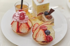 A variety of elegant desserts is arranged on a white plate. The selection includes pastries topped with strawberries, raspberries, blueberries, and apple slices. One dessert is adorned with pink whipped cream and a raspberry on top. In the background, a small glass contains an amber-colored drink.