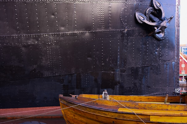 A large metal ship hull with riveted panels dominates the background, while in the foreground, a small wooden boat with a vibrant yellow hue is moored. The ship appears to have a black anchor attached at the side, with a rope extending from the boat.