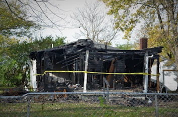 A severely burned and charred structure, likely a residential house, surrounded by vegetation and a metal fence. Yellow caution tape is stretched across the front, indicating restricted access. The roof is partially collapsed, and the walls are darkened due to fire damage. The sky is clear, and some trees in the background show green and yellow leaves.
