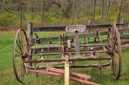 An old, rusty agricultural implement with large metal wheels and a metal seat is located in a grassy field. Behind it, a wooden fence and leafless trees suggest an autumn setting.