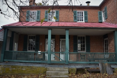 A two-story brick house features green shutters on its windows and a red metal roof. The front porch has teal railings and supports, with a set of concrete steps leading up to it. Fallen leaves cover the ground while bare branches from a nearby tree extend into view.