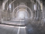 An interior of a large, vaulted space featuring repeated metal rods creating an optical illusion of depth. Sunlight filters through arched windows, casting intricate patterns of light and shadow on the walls and floor. At the far end, a large circular shape is visible, adding to the geometric complexity of the scene.