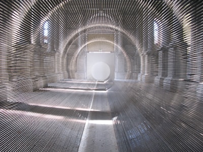 An interior of a large, vaulted space featuring repeated metal rods creating an optical illusion of depth. Sunlight filters through arched windows, casting intricate patterns of light and shadow on the walls and floor. At the far end, a large circular shape is visible, adding to the geometric complexity of the scene.
