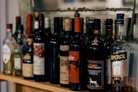 A collection of various liquor bottles is displayed on a wooden shelf. There are different types of alcoholic beverages including wine and spirits. The bottles have colorful labels and are placed in a row close to each other.