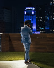 A person in a light blue suit is standing on a rooftop terrace at night, talking on the phone. The setting includes a wooden fence and a patch of artificial grass. In the background, a cityscape with a prominent building illuminated in blue lights creates an urban atmosphere.