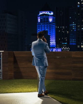 A person in a light blue suit is standing on a rooftop terrace at night, talking on the phone. The setting includes a wooden fence and a patch of artificial grass. In the background, a cityscape with a prominent building illuminated in blue lights creates an urban atmosphere.