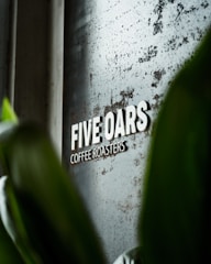 A metallic sign with the words 'FIVE OARS COFFEE ROASTERS' is mounted on a textured, slightly rusted surface. Green leaves partially obscure the view, suggesting a natural, organic ambiance.