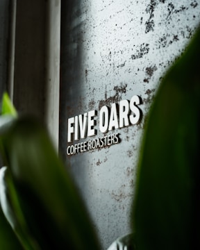 A metallic sign with the words 'FIVE OARS COFFEE ROASTERS' is mounted on a textured, slightly rusted surface. Green leaves partially obscure the view, suggesting a natural, organic ambiance.