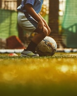 A young child wearing a blue jersey and white shorts is preparing to kick a soccer ball on a grassy field. The scene is warmly lit, suggesting a late afternoon or early evening environment.