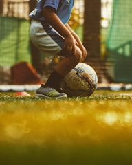 A young child wearing a blue jersey and white shorts is preparing to kick a soccer ball on a grassy field. The scene is warmly lit, suggesting a late afternoon or early evening environment.