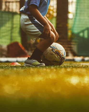 A young child wearing a blue jersey and white shorts is preparing to kick a soccer ball on a grassy field. The scene is warmly lit, suggesting a late afternoon or early evening environment.