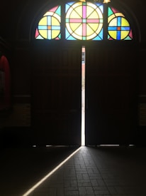 A dimly lit interior space features a set of large wooden doors, slightly ajar, allowing a beam of light to stream in and align perfectly along the tiled floor. Above the doors, a half-circle stained glass window showcases a vibrant geometric pattern with circles and squares in colors like blue, yellow, and red.