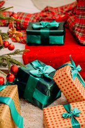 A collection of beautifully wrapped gifts in various patterns and colors are placed under a decorated Christmas tree. The presents have green and teal ribbons adorning them, and the gift wrap includes solid colors and polka dot patterns. The tree is adorned with red baubles and a gingerbread man ornament. A red and plaid fabric serves as a festive backdrop.