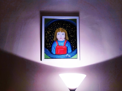 A framed painting of a child with blonde hair wearing a blue shirt and a red overall is centrally mounted on a wall. The image appears to be illuminated by soft light from a lamp at the bottom center, creating a warm and intimate atmosphere.