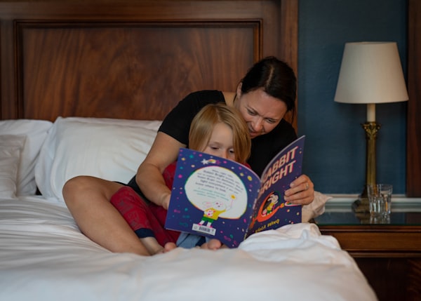 a little blonde girl with red pajamas in bed reading a children book with her mother