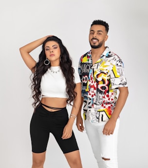 A woman and a man are posing against a plain backdrop. The woman has long, wavy hair and wears a white crop top with black shorts, accessorized with large hoop earrings and a necklace. The man sports a colorful, patterned shirt with white pants, and has short hair and a beard. They both have a confident stance.