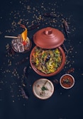 A top-down view of a traditional Indian meal featuring a clay pot filled with a vibrant, colorful rice dish garnished with fresh herbs and spices. Accompanying this are two small bowls containing sauces or condiments, one white and creamy, the other red and spicy. A glass of cocktail with ice, orange slice, and a cherry garnish is visible alongside scattered dry red chilies and seeds on a dark surface, enhancing the rustic aesthetic.