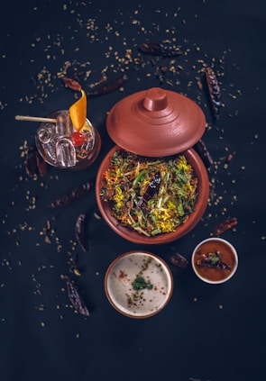A top-down view of a traditional Indian meal featuring a clay pot filled with a vibrant, colorful rice dish garnished with fresh herbs and spices. Accompanying this are two small bowls containing sauces or condiments, one white and creamy, the other red and spicy. A glass of cocktail with ice, orange slice, and a cherry garnish is visible alongside scattered dry red chilies and seeds on a dark surface, enhancing the rustic aesthetic.