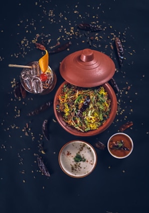 A top-down view of a traditional Indian meal featuring a clay pot filled with a vibrant, colorful rice dish garnished with fresh herbs and spices. Accompanying this are two small bowls containing sauces or condiments, one white and creamy, the other red and spicy. A glass of cocktail with ice, orange slice, and a cherry garnish is visible alongside scattered dry red chilies and seeds on a dark surface, enhancing the rustic aesthetic.