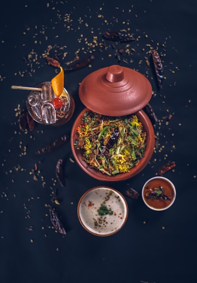 A top-down view of a traditional Indian meal featuring a clay pot filled with a vibrant, colorful rice dish garnished with fresh herbs and spices. Accompanying this are two small bowls containing sauces or condiments, one white and creamy, the other red and spicy. A glass of cocktail with ice, orange slice, and a cherry garnish is visible alongside scattered dry red chilies and seeds on a dark surface, enhancing the rustic aesthetic.