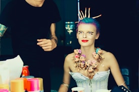 A person with colorful hair and elaborate makeup sits in a preparation area. They are adorned with a unique necklace made of multiple doll heads with varying expressions. The background features someone standing with artistic tattoos on their arm, possibly assisting in the preparation. Bright containers, possibly of makeup or styling products, are scattered on the table in the foreground.