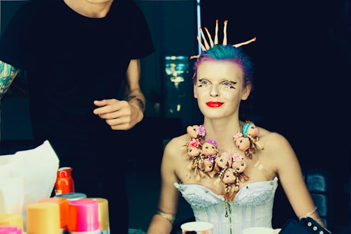 A person with colorful hair and elaborate makeup sits in a preparation area. They are adorned with a unique necklace made of multiple doll heads with varying expressions. The background features someone standing with artistic tattoos on their arm, possibly assisting in the preparation. Bright containers, possibly of makeup or styling products, are scattered on the table in the foreground.