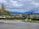Residential neighborhood with modern multi-story houses surrounded by trees. A paved road and some street lamps are visible in the foreground. The background features a range of mountains under a cloudy sky.