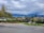 Residential neighborhood with modern multi-story houses surrounded by trees. A paved road and some street lamps are visible in the foreground. The background features a range of mountains under a cloudy sky.