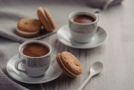 Two cups of coffee are placed on saucers with a creamy texture and earthy tones. They are accompanied by sandwich cookies with a layer of cream, set on a light wooden surface. A cloth napkin is visible in the background, adding a cozy touch to the scene.