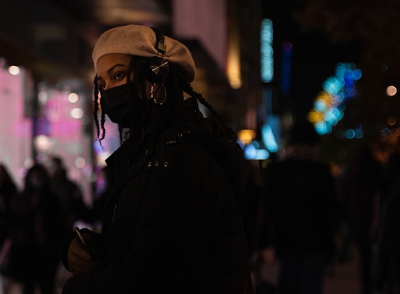 A person wearing a light-colored beret and headphones stands in an urban setting at night. The dim lighting and colorful bokeh in the background suggest a busy city street. The individual has braided hair and is wearing a dark jacket while holding a smartphone.