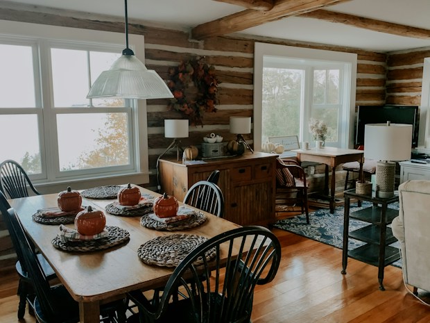 A cozy, rustic dining room with a wooden table set for six. The table is adorned with autumn-themed decorations, including small orange pumpkin ornaments on woven placemats. Large windows allow natural light to illuminate the space, highlighting the wooden beams and paneled walls. A sideboard holds a few decorative items, and there is a hint of greenery visible through the windows.
