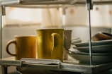 A close-up view of a kitchen dish rack holding several items, including two colorful mugs and a stack of neatly arranged plates. The mugs are yellow, and the plates are mostly white. Soft, natural light filters through a nearby window, adding a warm ambiance to the scene.
