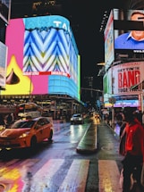 A vibrant urban scene at night featuring bright, colorful billboards and signs with people walking along the sidewalk. The wet streets reflect the lights of yellow taxis and surrounding buildings. Advertising for shows and chocolate brands is prominently displayed.