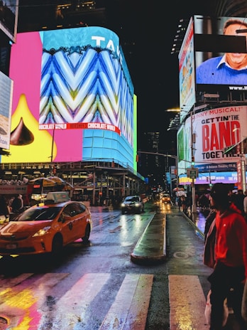 A vibrant urban scene at night featuring bright, colorful billboards and signs with people walking along the sidewalk. The wet streets reflect the lights of yellow taxis and surrounding buildings. Advertising for shows and chocolate brands is prominently displayed.