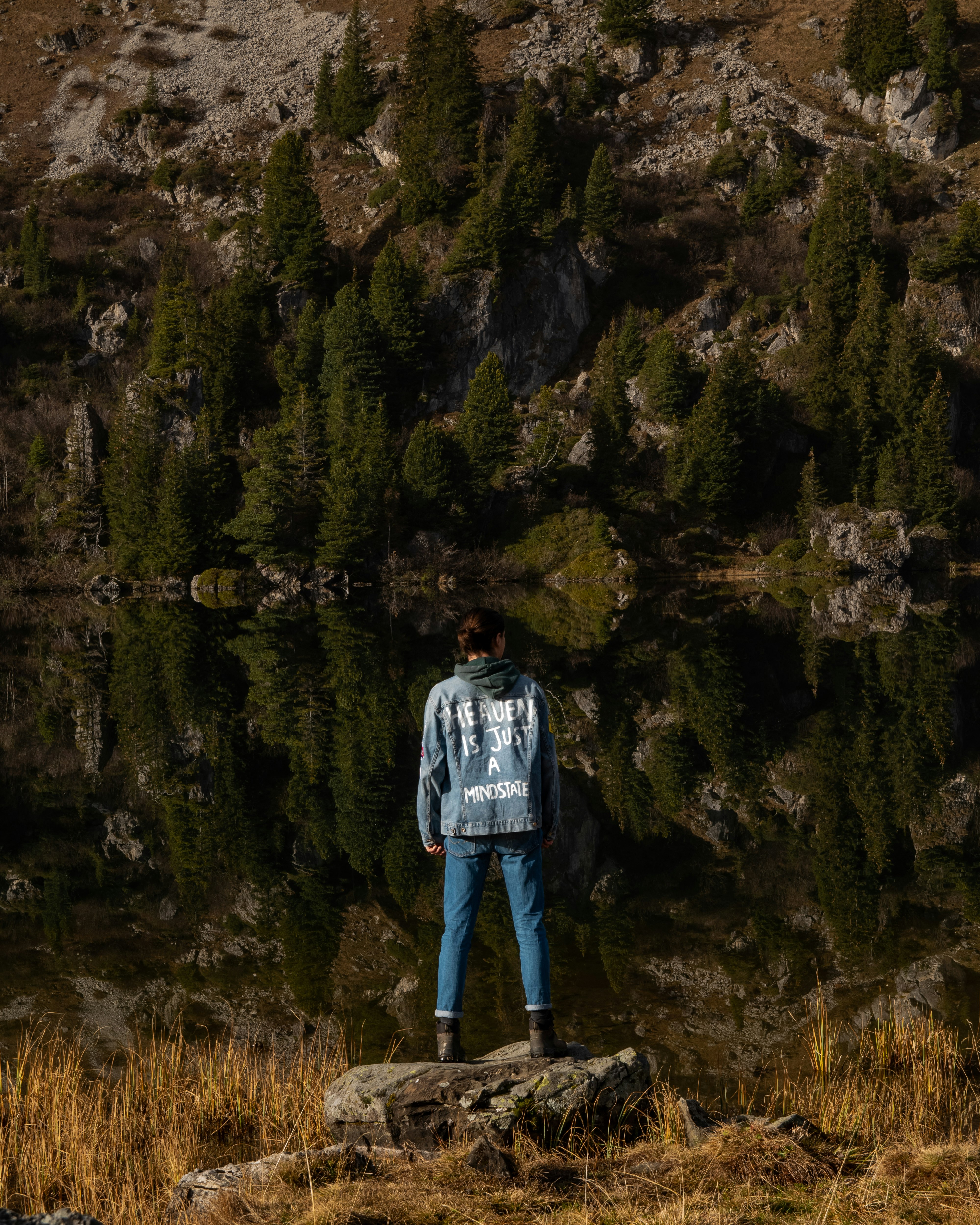 man in blue jacket standing on rock near lake during daytime