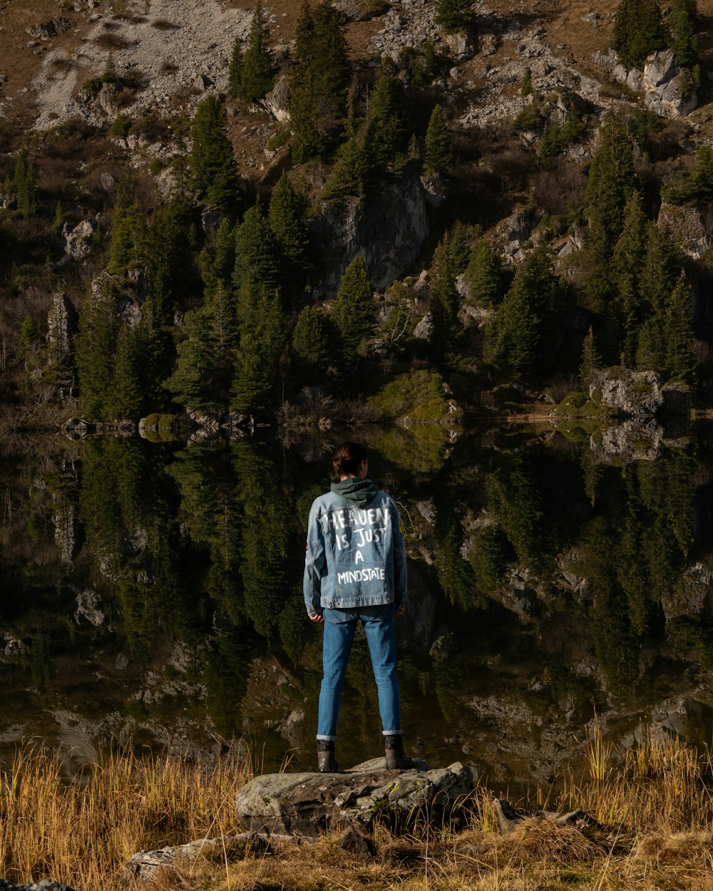 man in blue jacket standing on rock near lake during daytime