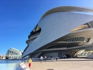 A futuristic architectural structure with sweeping curves and intricate lines dominates the scene. The building features large windows and terraces and is made of concrete and glass. In the foreground, a bright blue sky stretches overhead, and several people walk along a wide promenade near a pool of water.